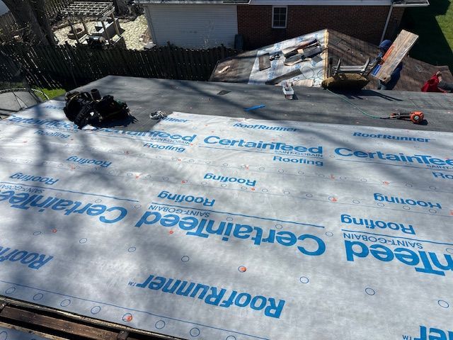 Roof under construction with white underlayment and blue printed logos, plus lumber and tools nearby