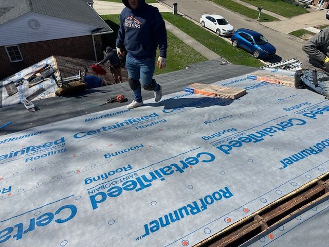 Worker standing on a roof underlayment during roof repair, with shingles and rooftop tools nearby.