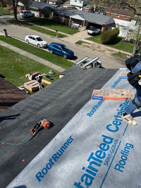A high-angle view of a residential roof with brown asphalt shingles against a backdrop of trees and a blue sky.