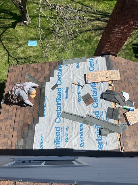 A close-up view of asphalt shingles on a roof meeting a white exterior wall corner above a gutter and dark decking.