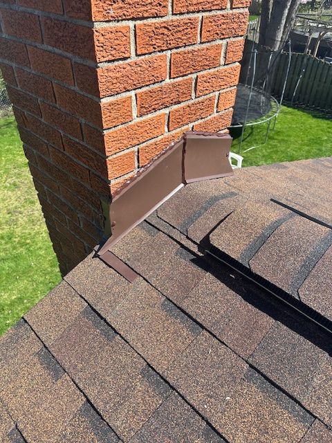 A metal vent pipe extending from a shingled roof, with a white chimney and green trees in the background.