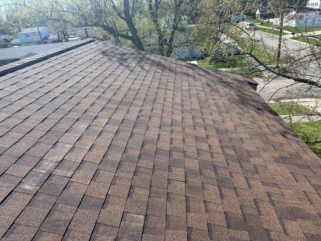 High-angle view of a brown shingled roof with two vent pipes, overlooking a neighborhood street and surrounding trees.