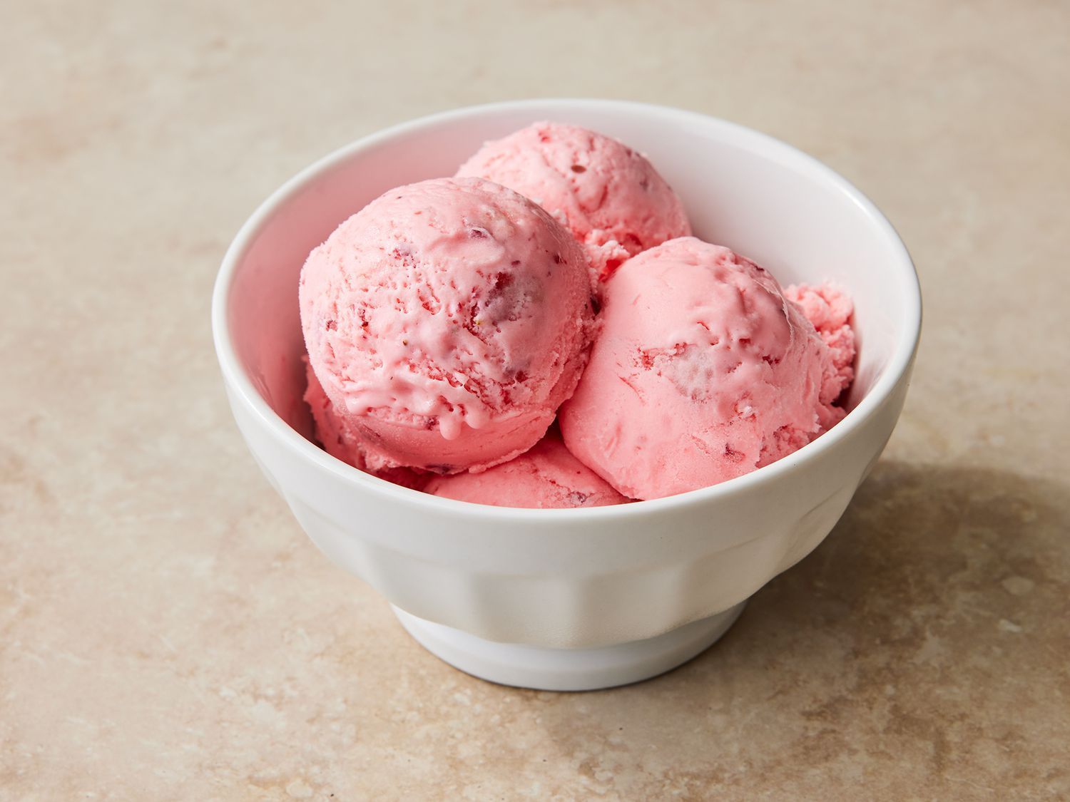A white bowl filled with four scoops of pink strawberry ice cream on a light stone surface.