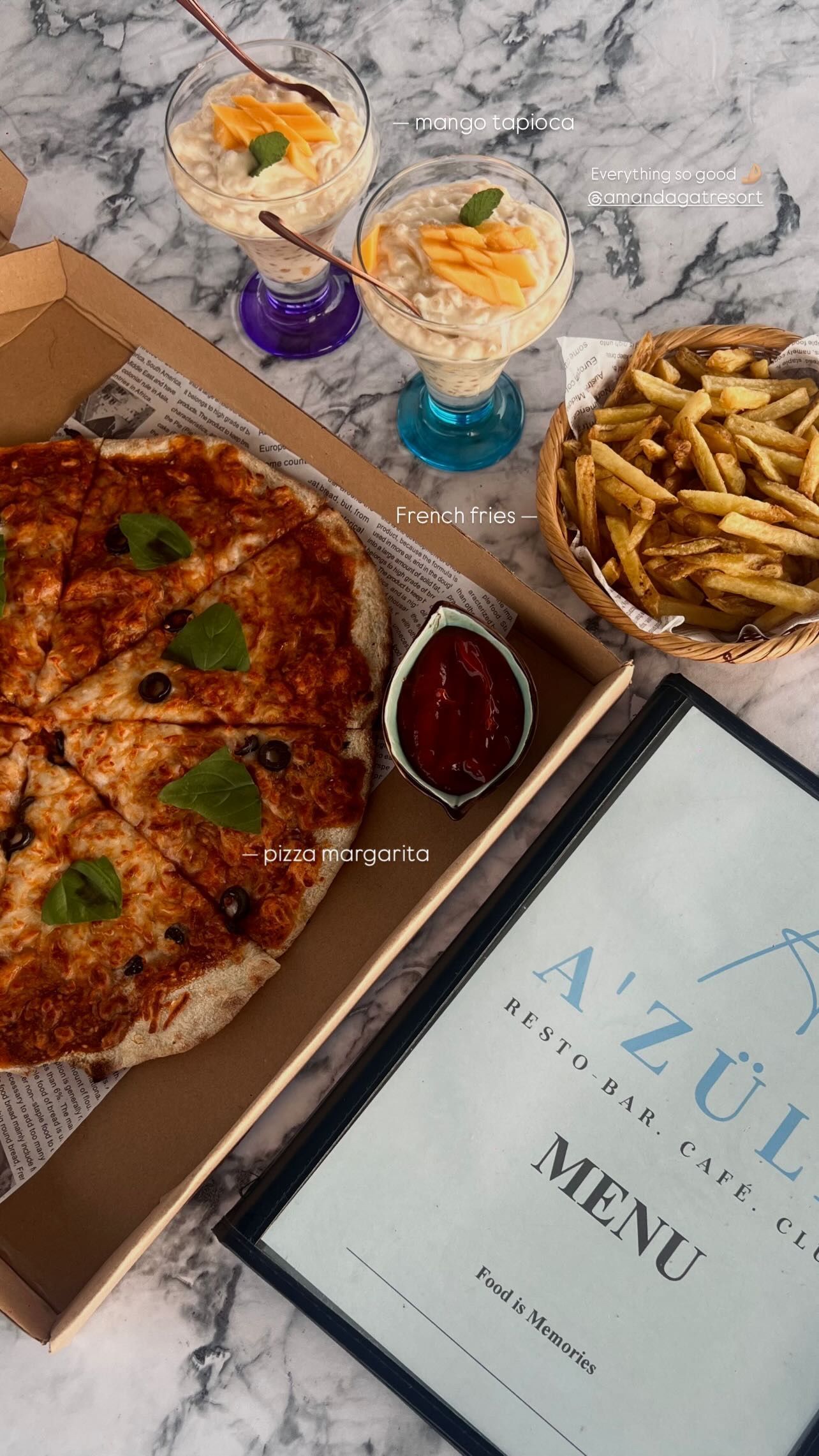 A pizza in a box, French fries, two fruit desserts, and an A'Zula menu on a marble table.