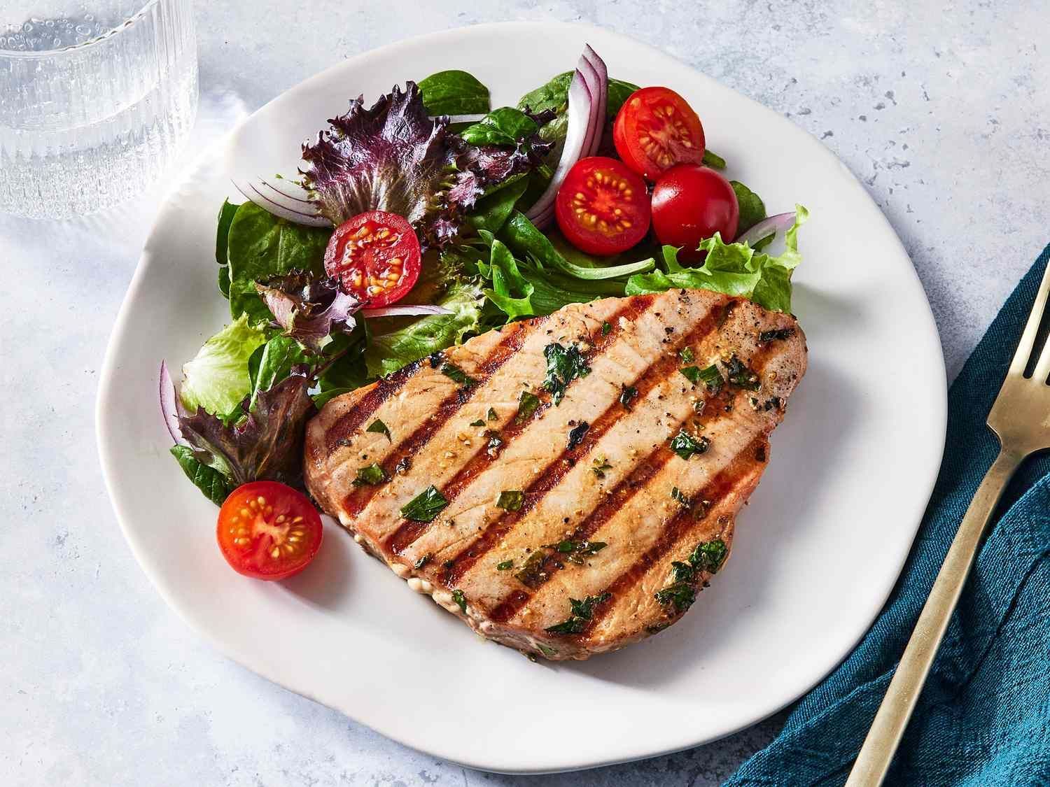 A grilled tuna steak seasoned with herbs, served on a white plate with a side salad of mixed greens, onion, and tomatoes.