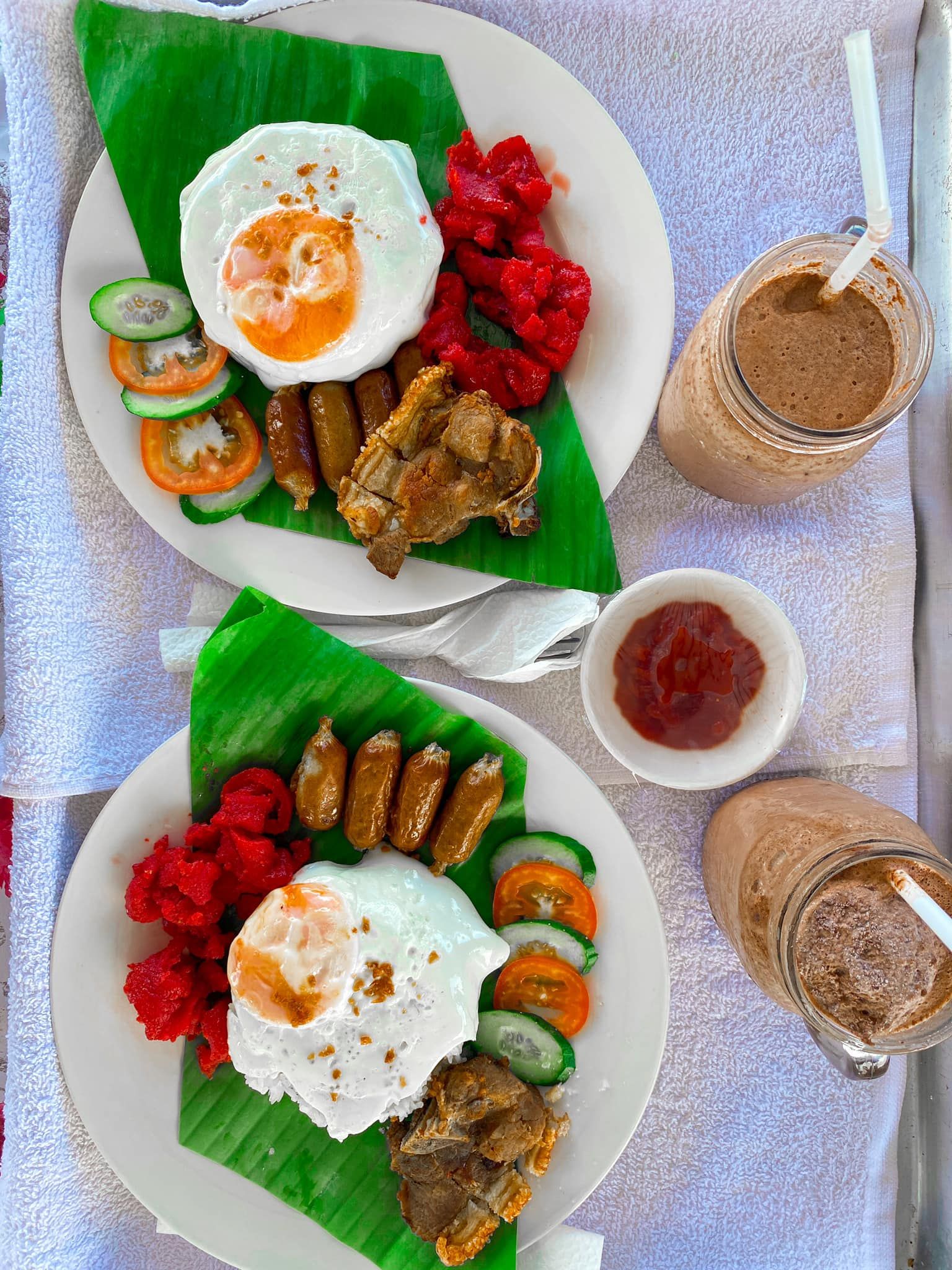 Two plates of breakfast with fried eggs, sausage, and red rice; smoothies and sauce on a table.