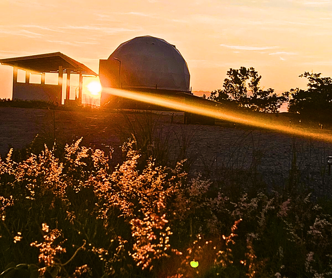 Sunset illuminates an observatory dome and shed. Beam of light shines across foreground of grass.