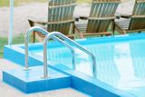 Pool with stainless steel ladder, blue water and surround, wooden chairs in background.