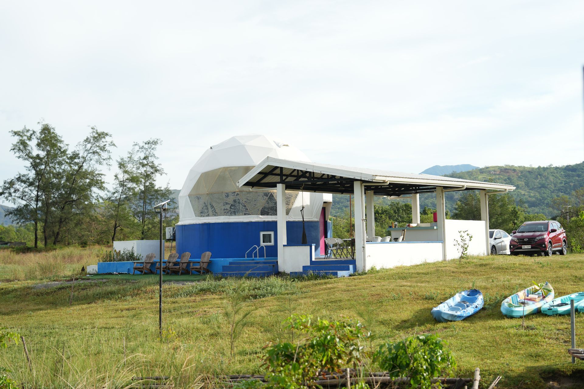 Blue and white geodesic dome building with open-air bar, surrounded by grassy field, boats, and a red car.