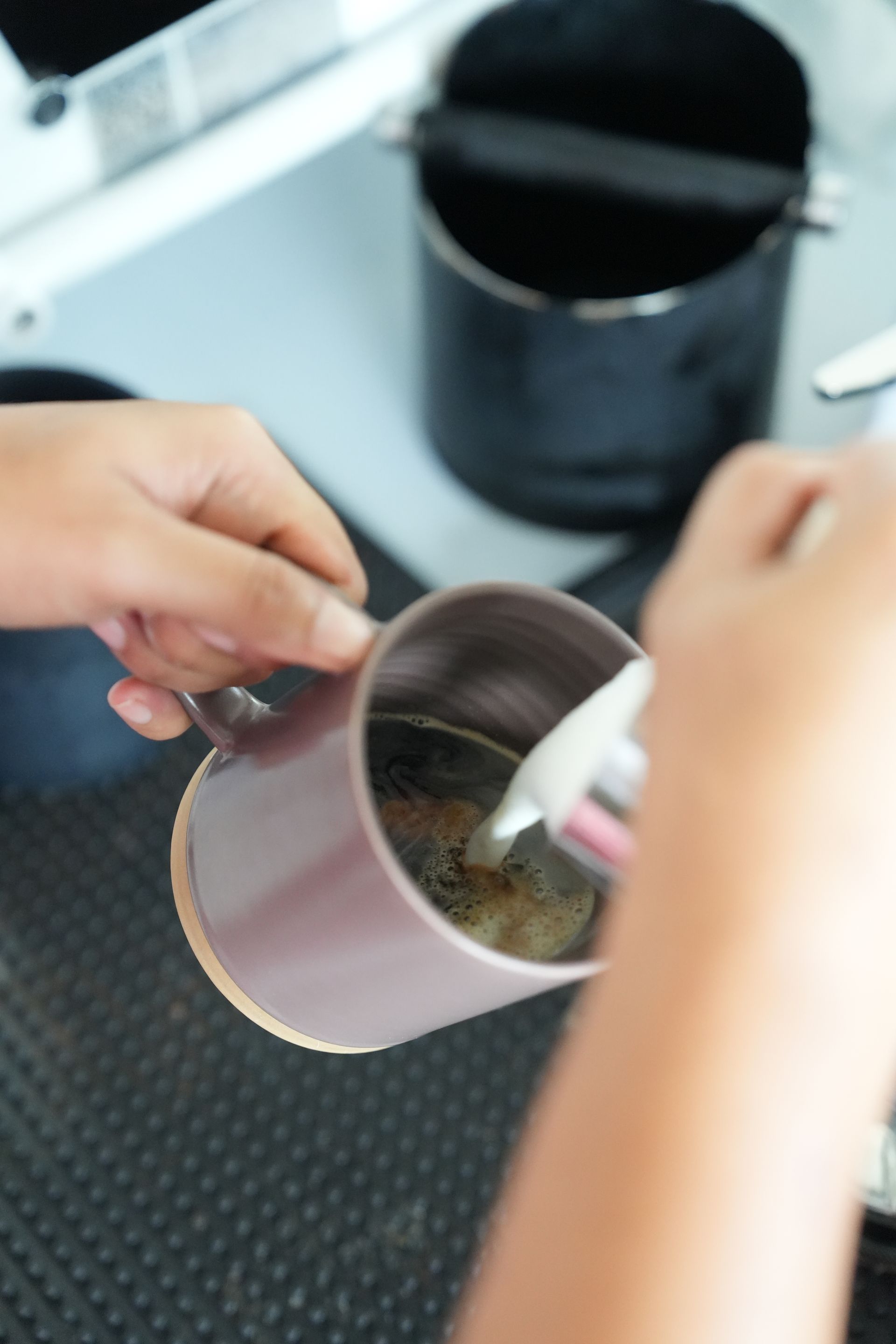 Person pouring steamed milk into coffee in a metal mug, near a coffee machine.