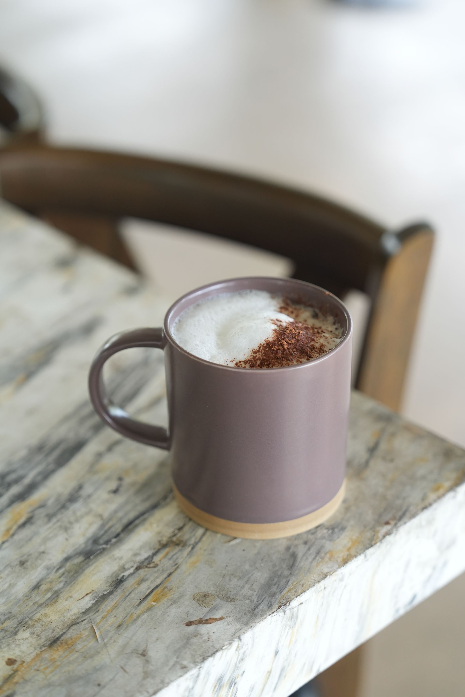 Mug of coffee with foam and sprinkles on a marble tabletop with a blurred wooden chair in the background.