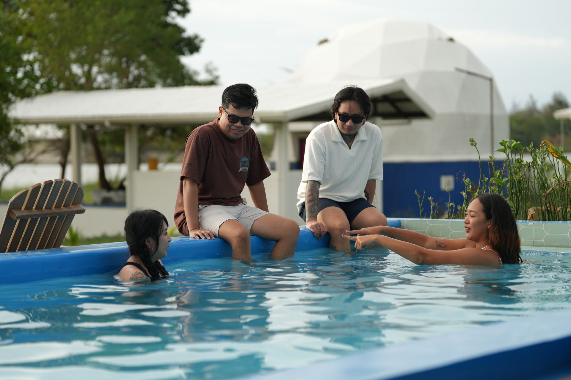 Two people sit on a pool edge as two others interact in the water; a white domed building is in the background.