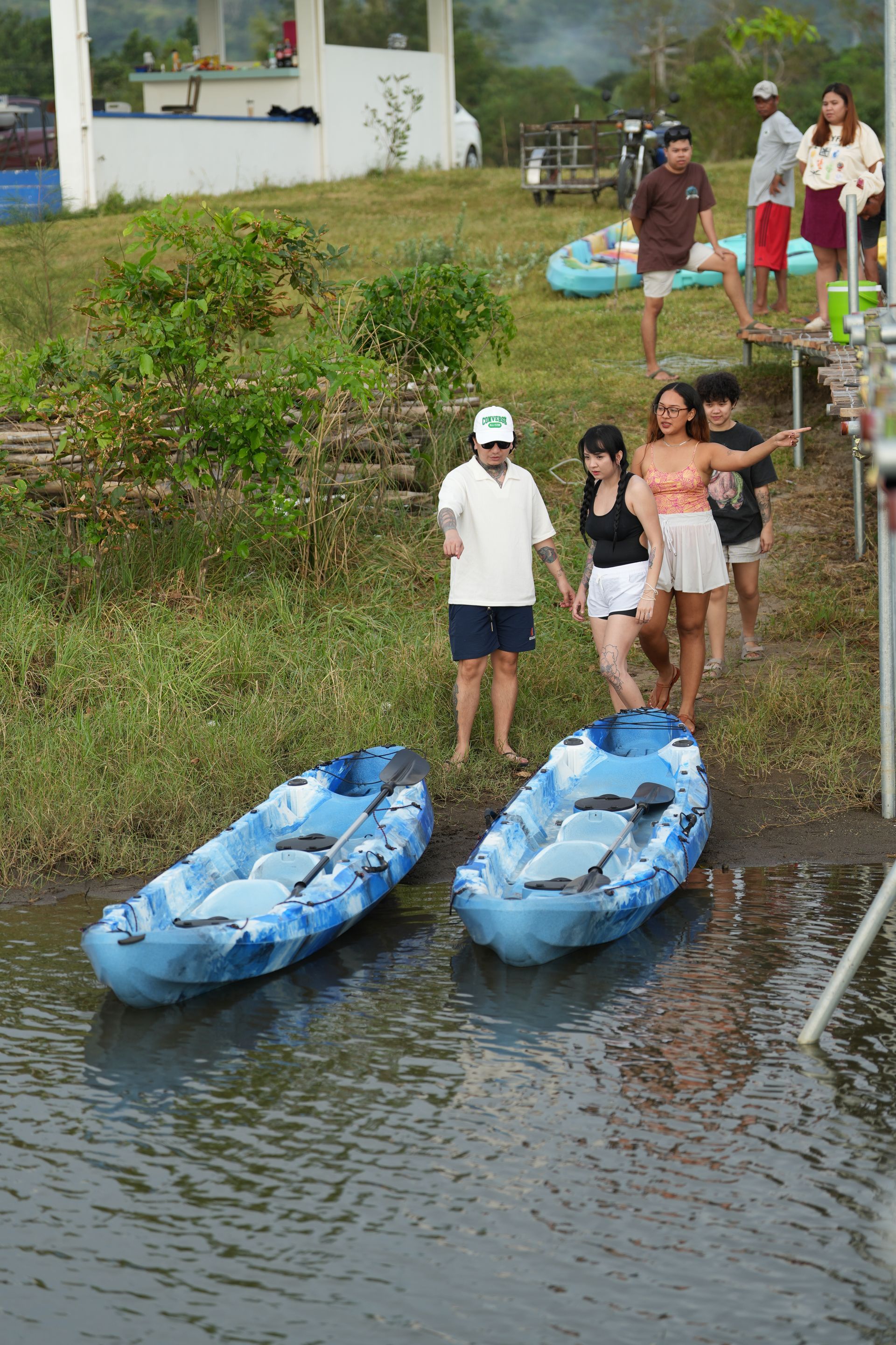 Two blue kayaks on water's edge. People stand nearby, some near a small building with a pier.