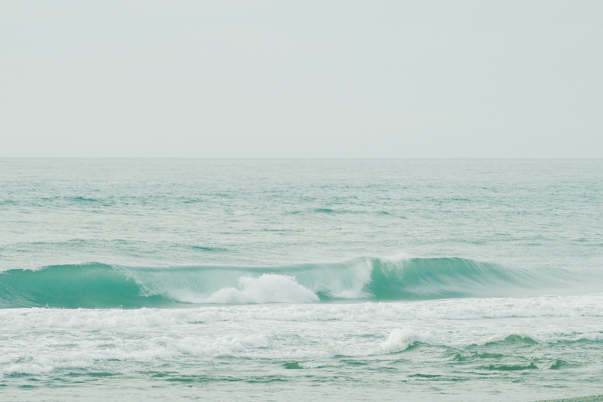Turquoise ocean waves rolling towards the shore under a hazy sky.