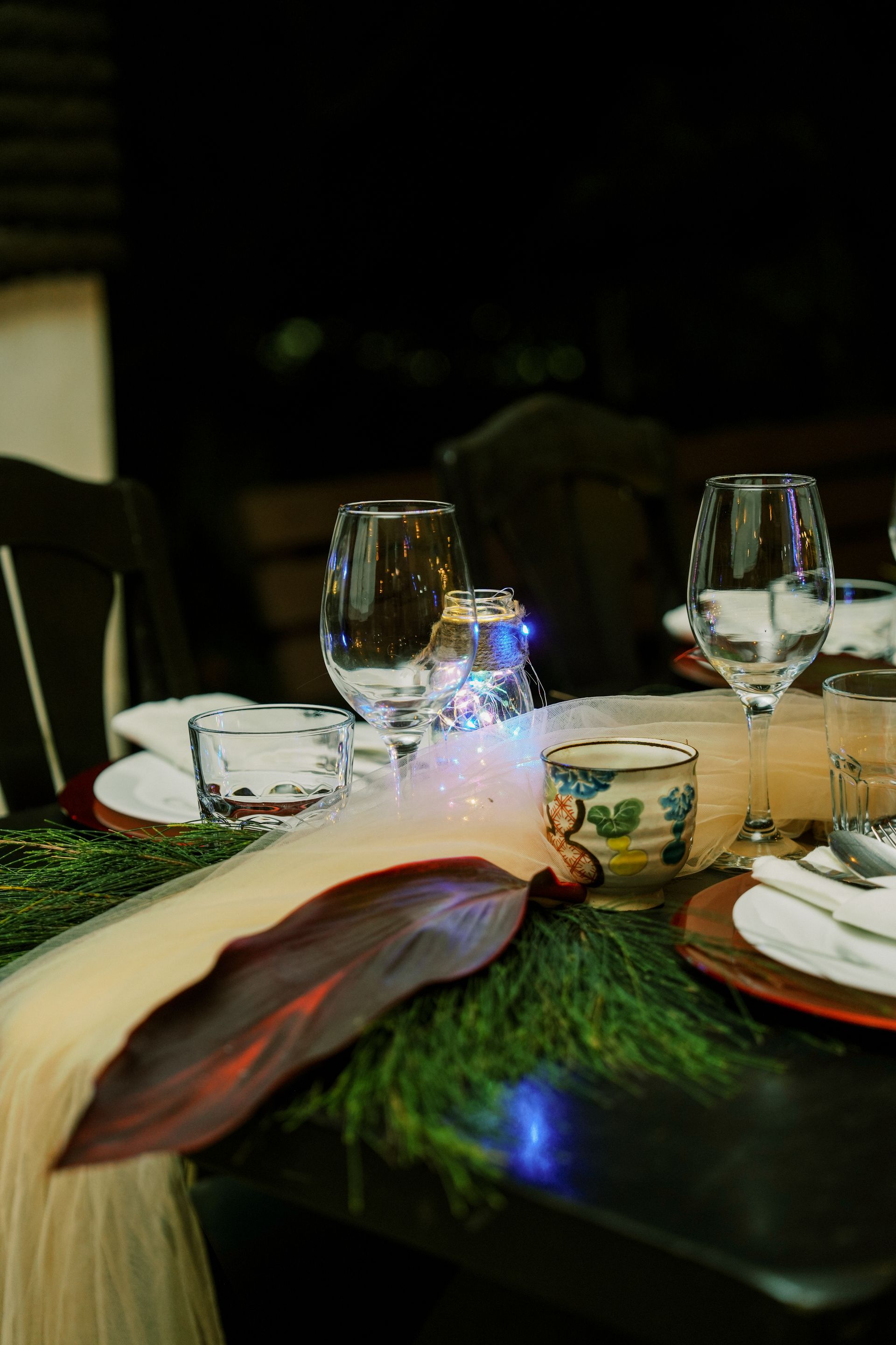 Formal table setting with glasses, plates, and a centerpiece, dimly lit in a dark room.