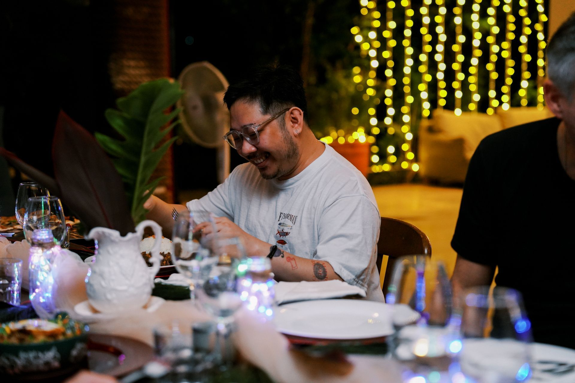 Man at a table, laughing. Lit table setting with other people; string lights in background.