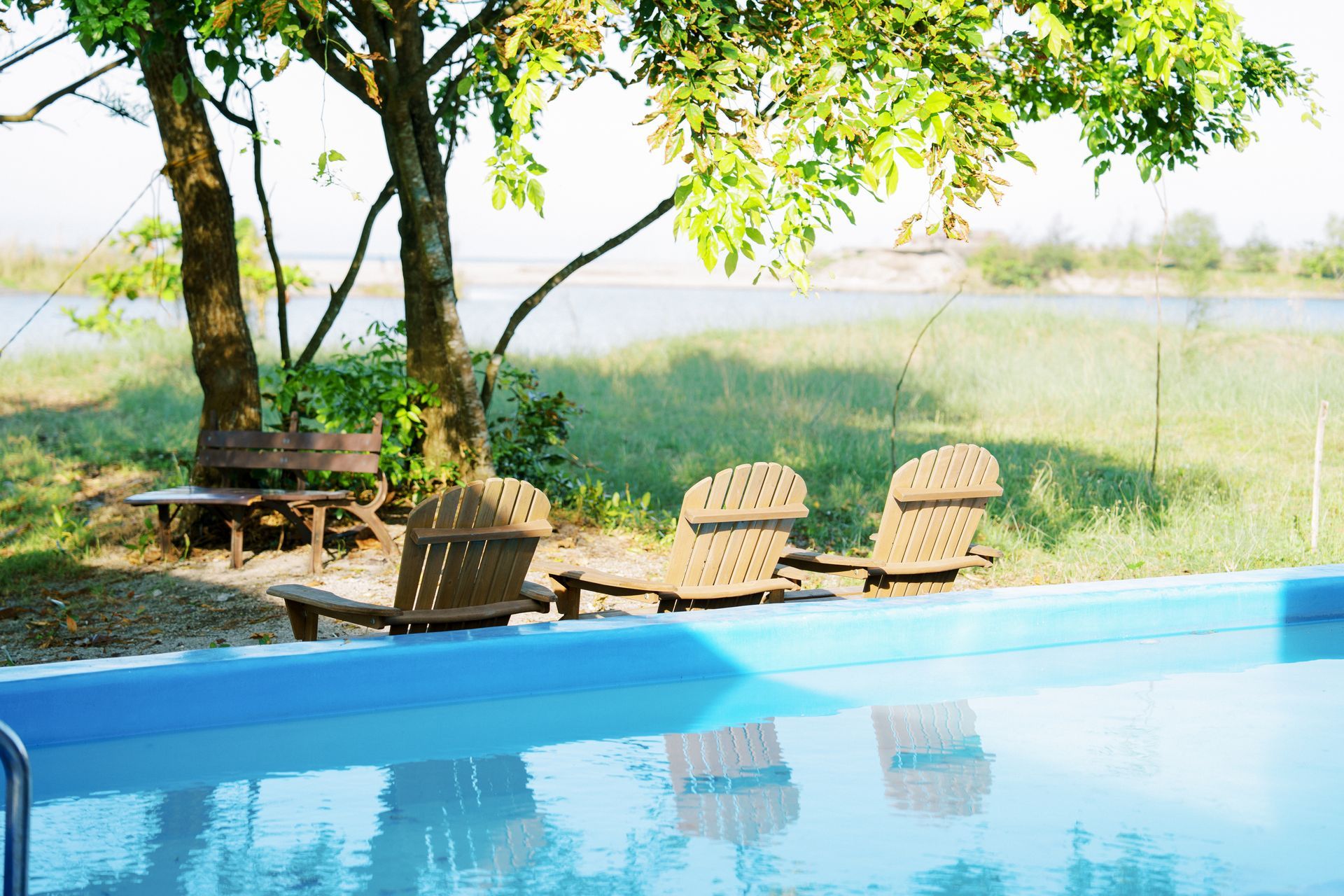 Swimming pool overlooking a grassy area with trees and a body of water. Wooden chairs and bench are nearby.