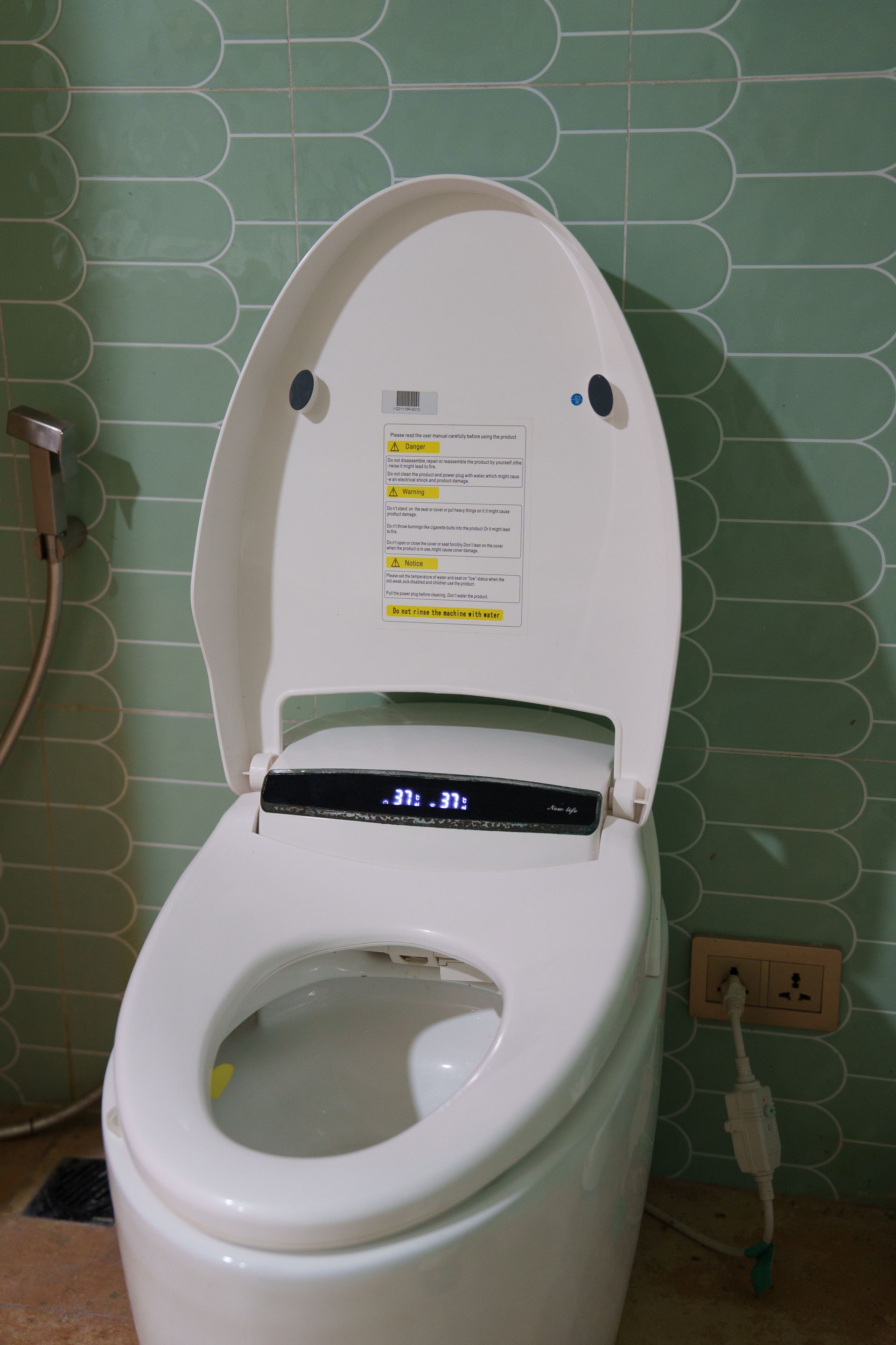 White, modern toilet with the lid up, controls visible. Set against green tiled wall, electrical outlet nearby.