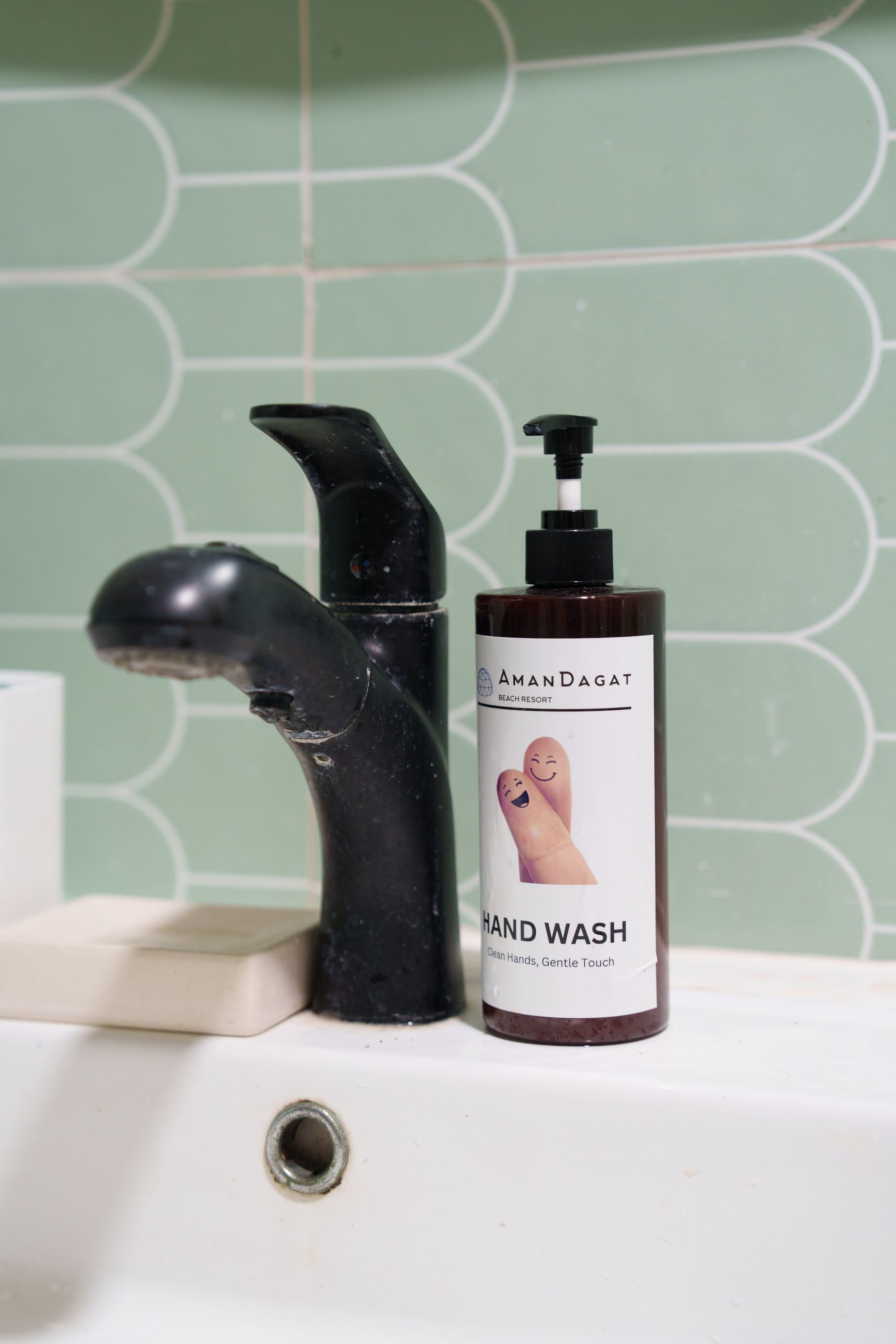 Black faucet and soap dispenser on a white sink, set against green tiles.