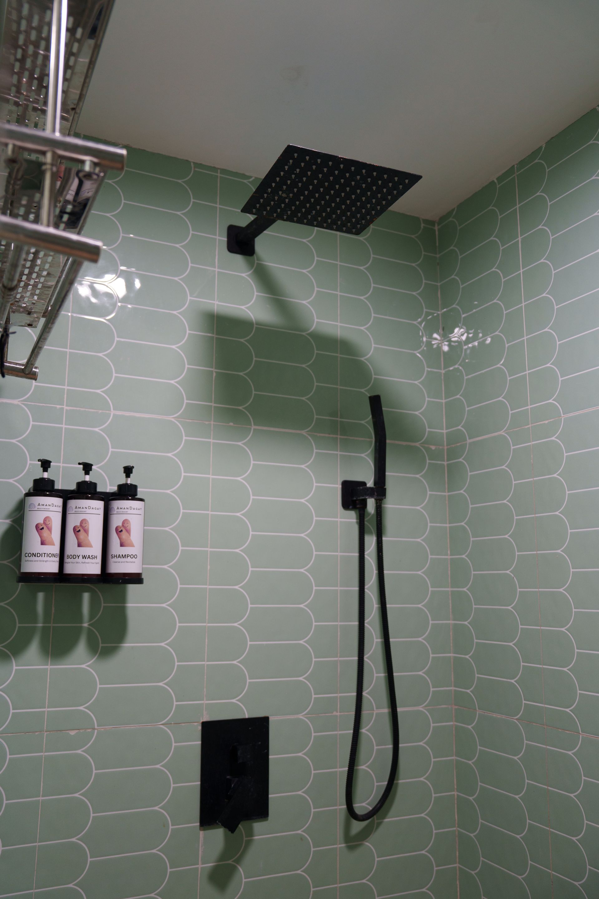 Black showerhead and handheld shower in a mint green tiled shower stall. Soap dispensers on the left.
