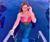 Woman in pink seashell bikini and sunglasses in blue water, exiting a pool by a ladder.
