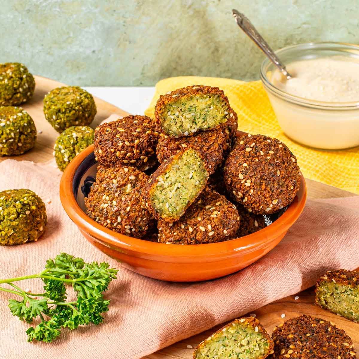 A bowl of crispy falafel on a pink napkin with a small bowl of dipping sauce and fresh parsley on a wooden surface.