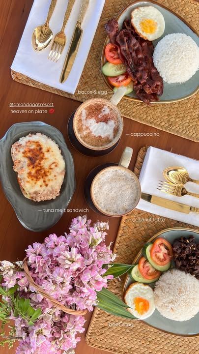 A flat-lay view of breakfast plates, coffee, and a bouquet of pink flowers on a wooden table with gold cutlery.