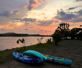 Two kayaks rest on shore, lake and sunset in the background.