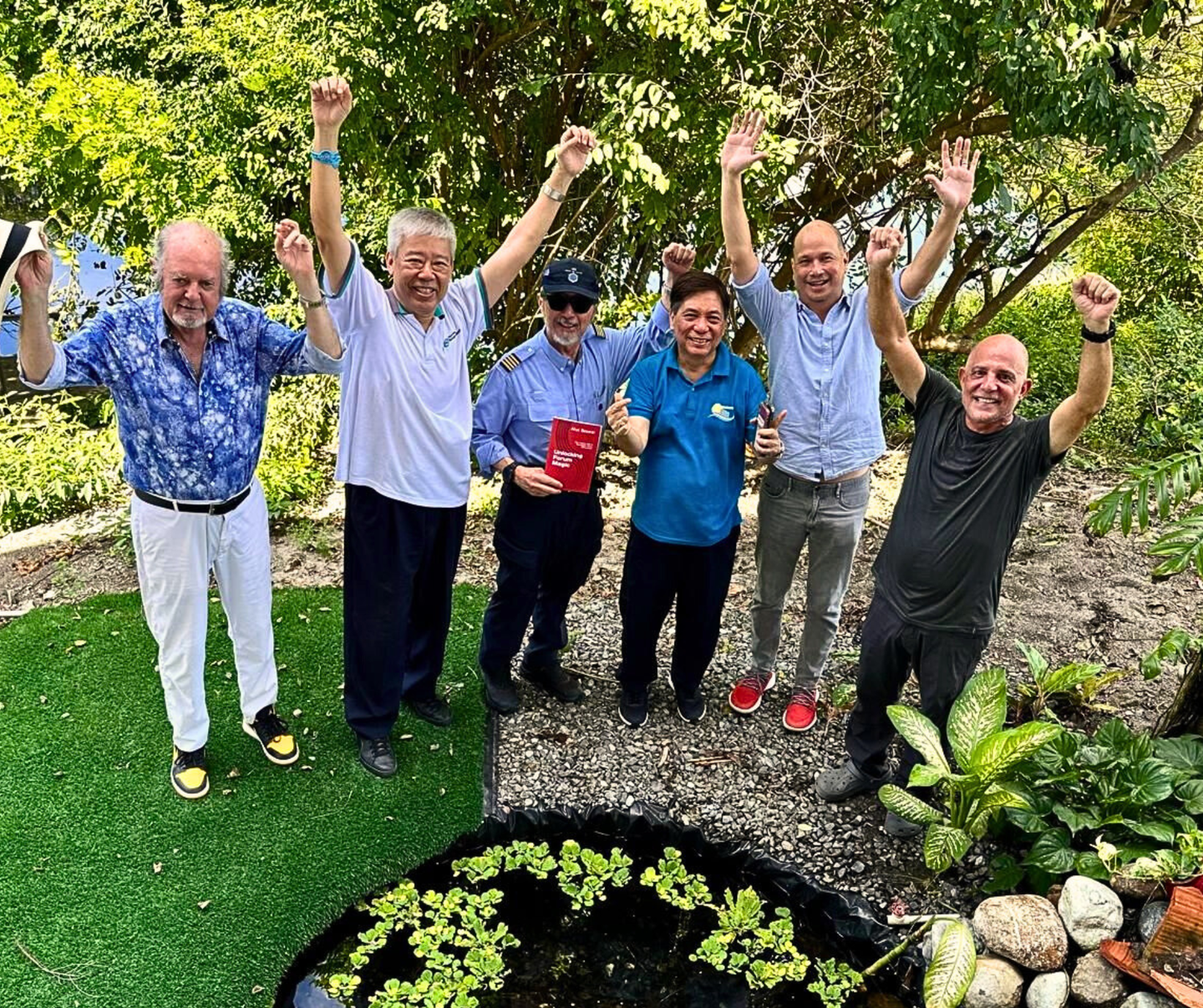 Group of six men with arms raised, celebrating near a pond, green foliage, bright sunny day.