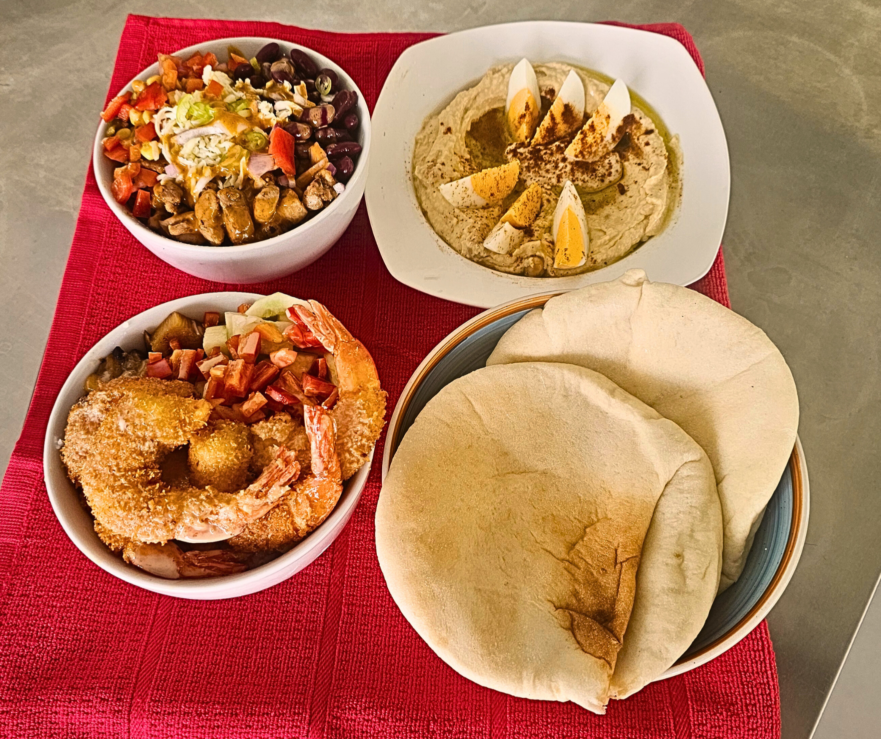 Overhead shot of a meal with hummus, pita bread, and bowls of food on a red tablecloth.