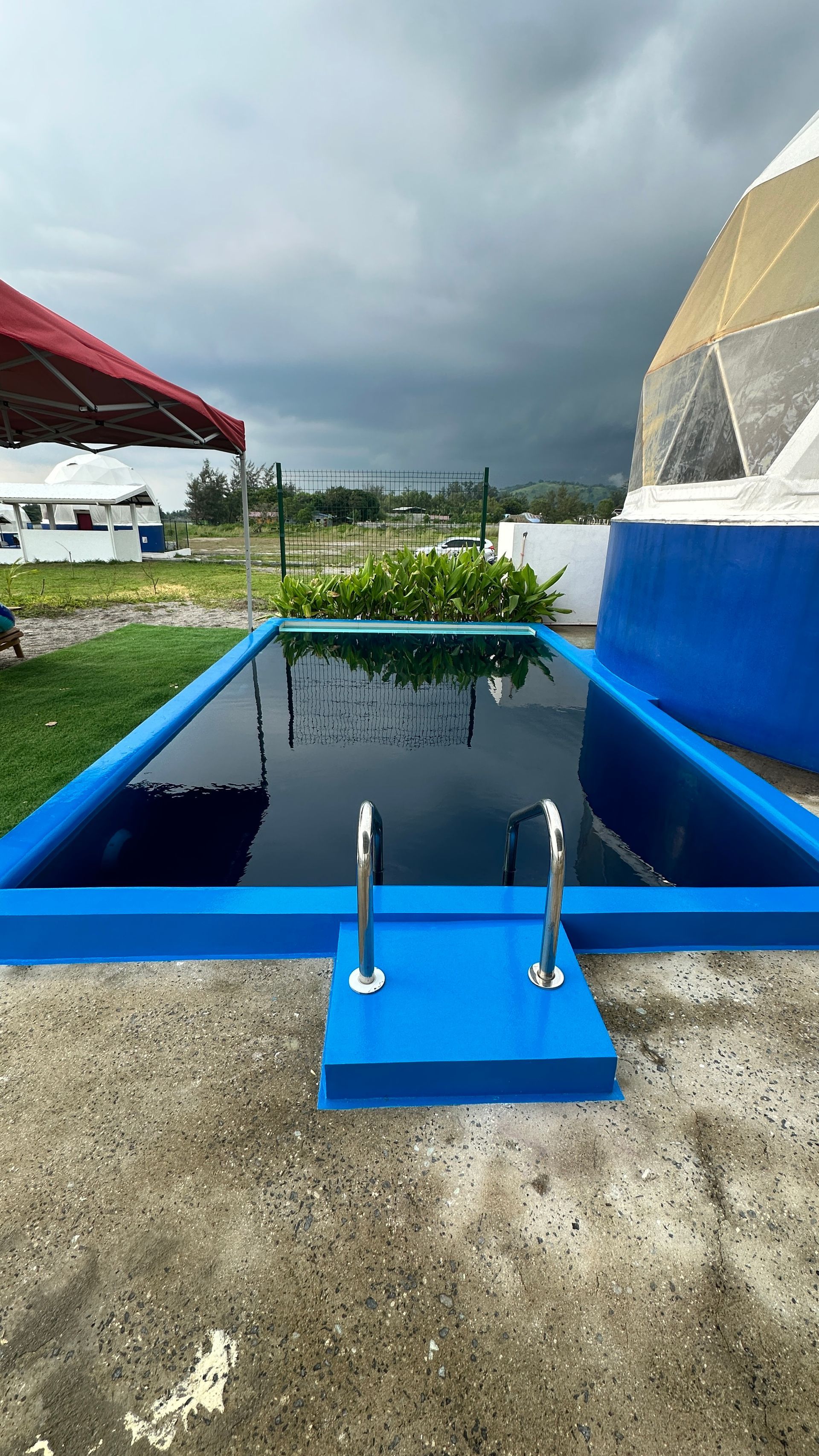 A blue rectangular pool with metal handrails on concrete, under a cloudy sky, next to a dome structure.