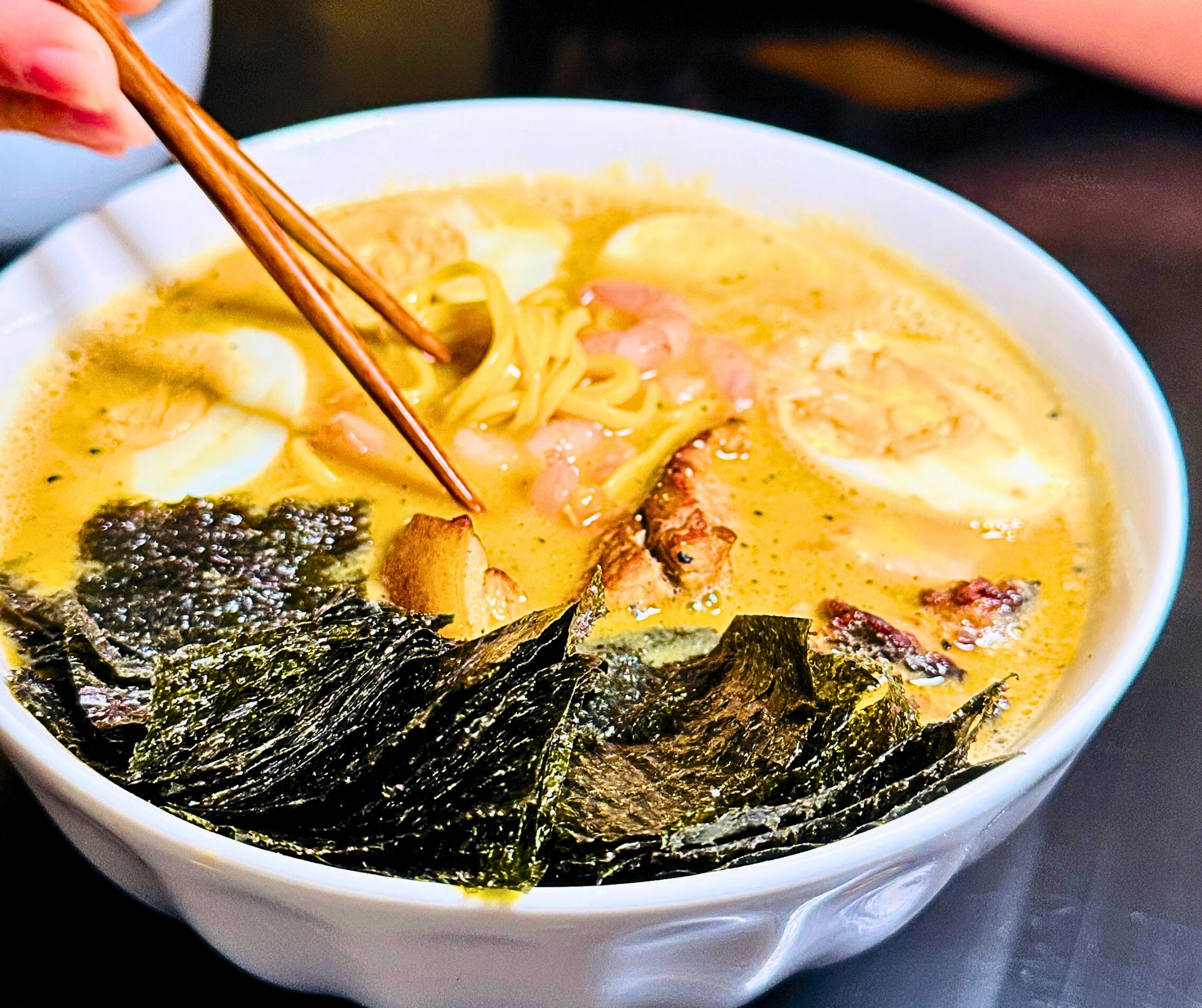 Ramen bowl with noodles, egg, seaweed, and chopsticks picking up noodles in a yellow broth.