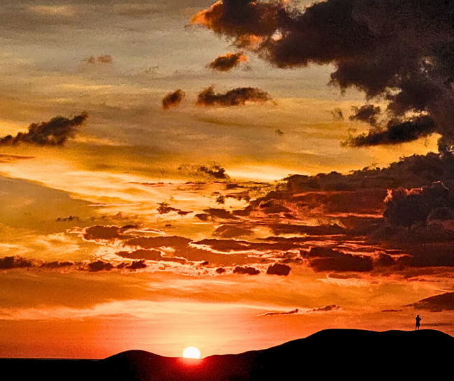 Sunset over dark hills, brilliant orange sky with scattered clouds, silhouetted figure.