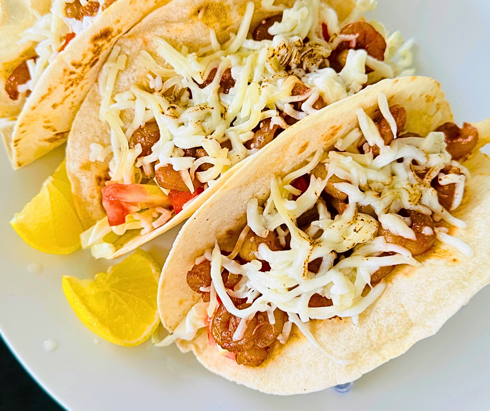 Tacos on a white plate with shredded cheese, tomato, and lemon slices.