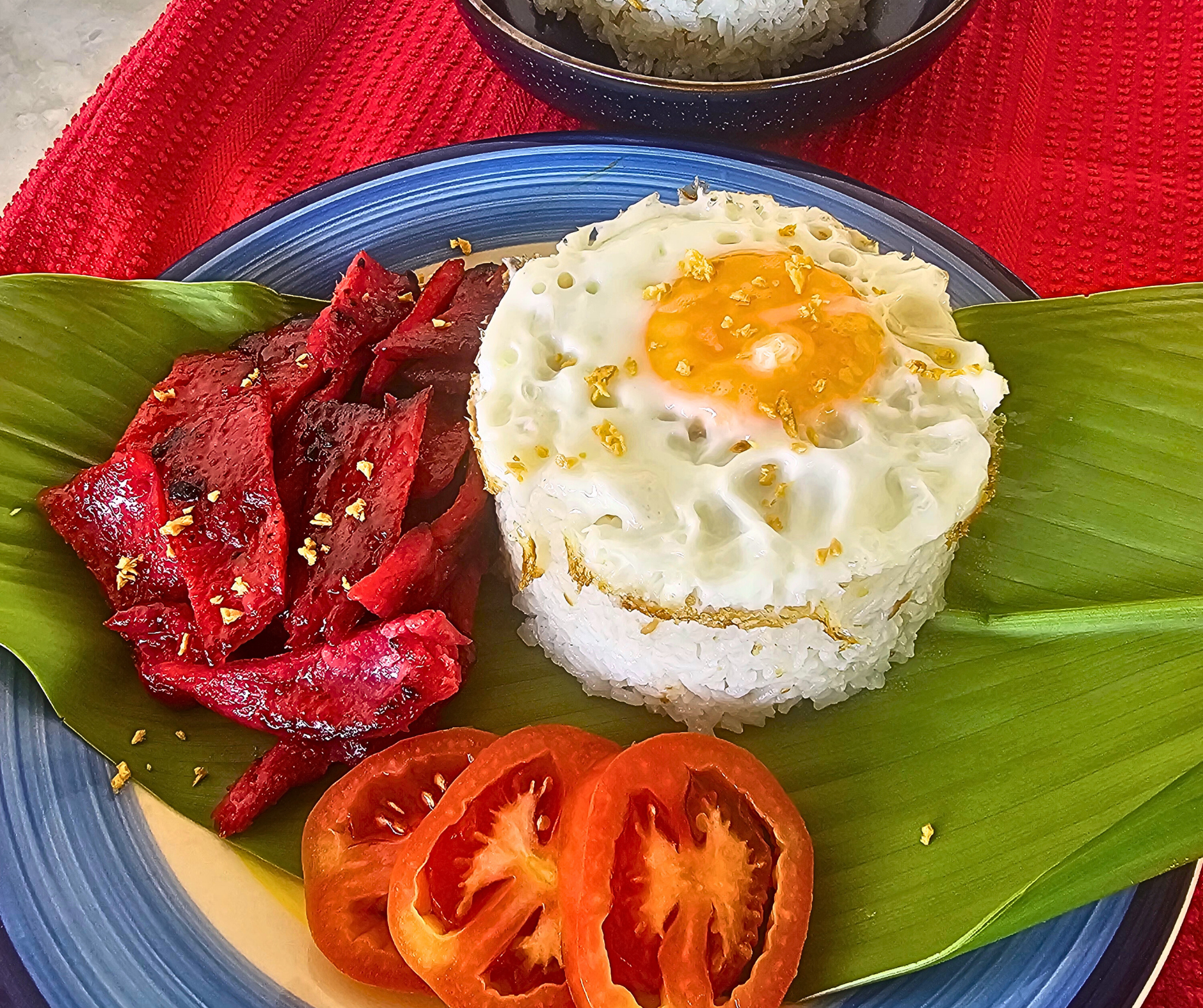 Filipino silog dish: rice, fried egg, red-colored meat, tomato slices on a green leaf.