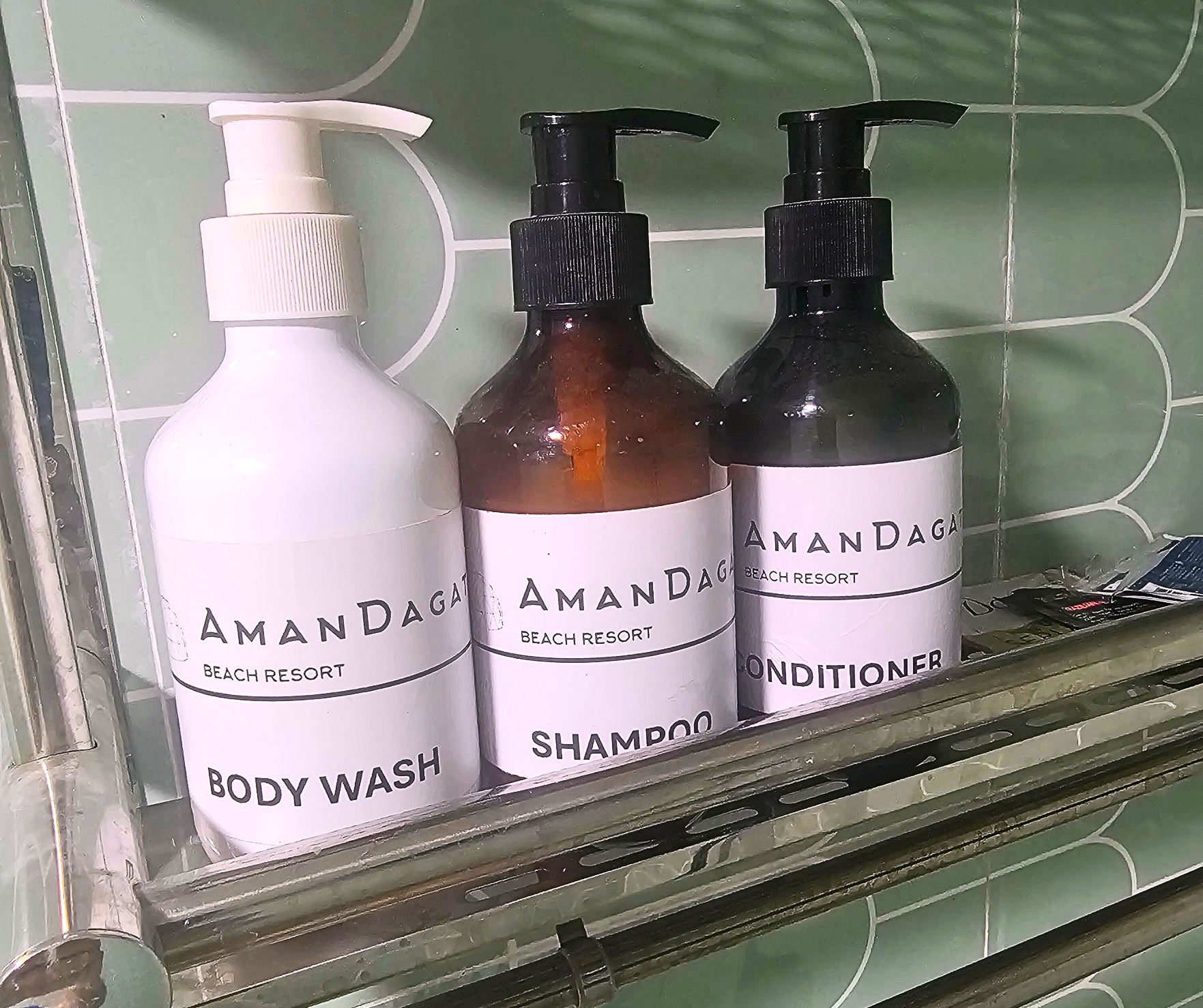 Three labeled pump bottles: body wash, shampoo, and conditioner on a metal shelf in a bathroom.