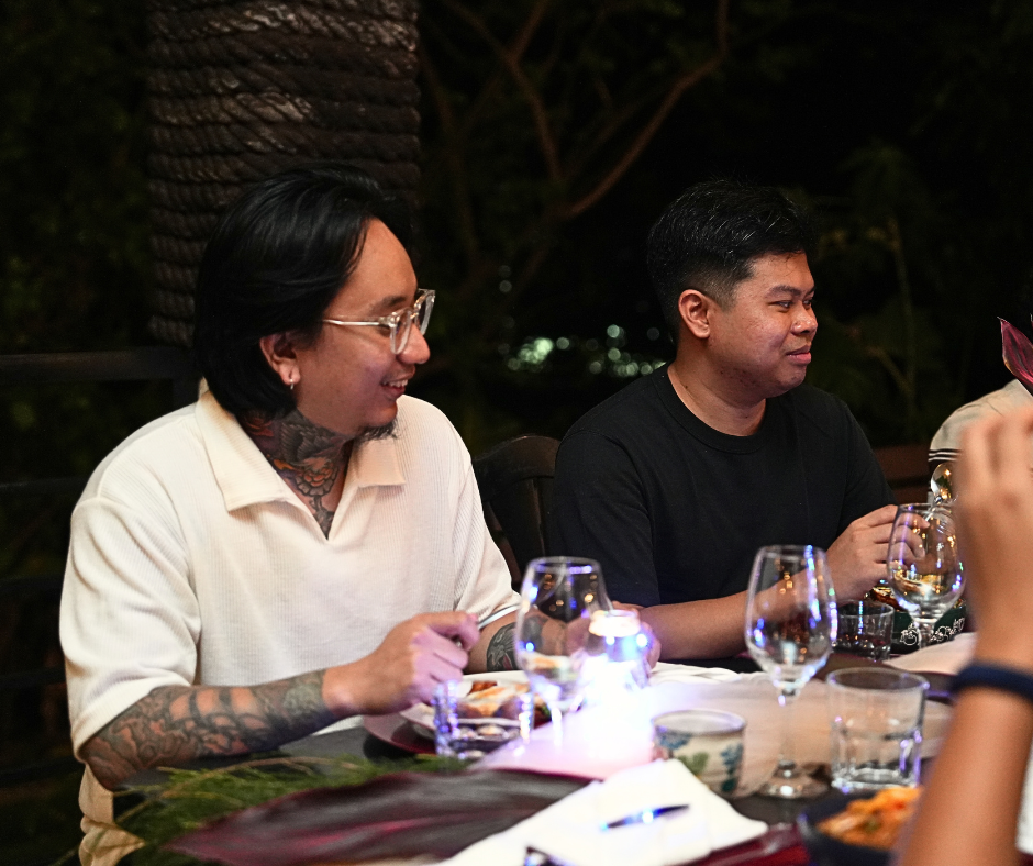 Two men at a table, eating and talking. One with tattoos and glasses, the other in a black shirt, at an outdoor dinner.