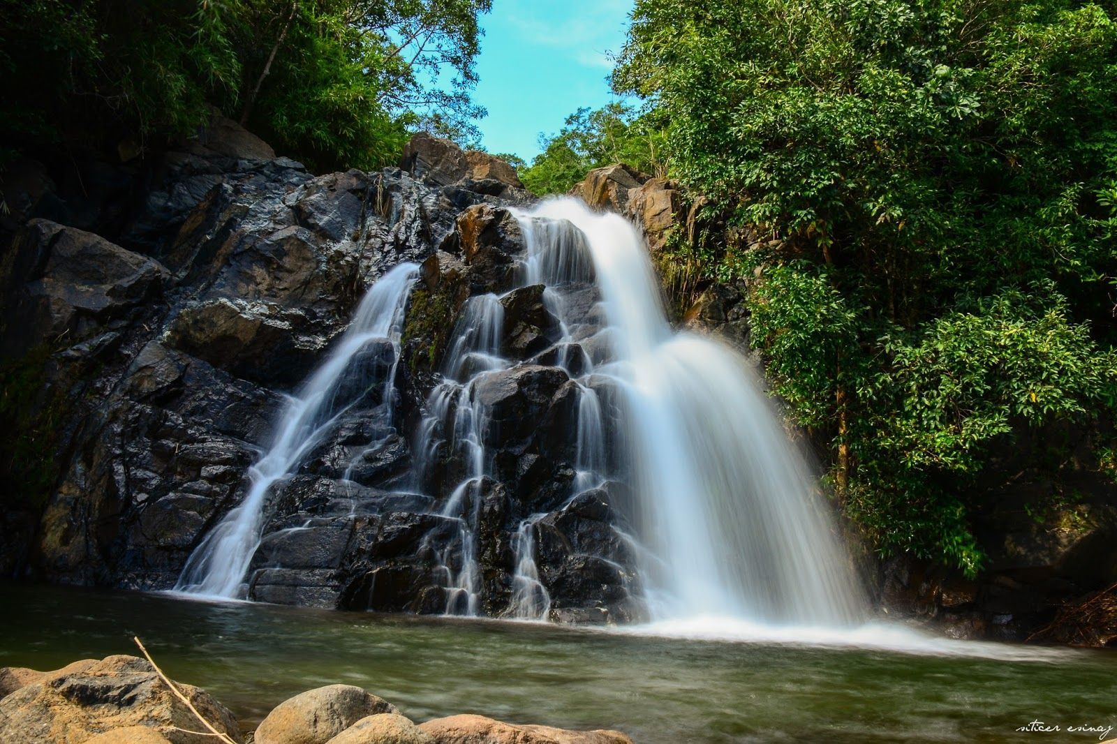 Waterfall cascading over dark rocks into a pool, surrounded by lush green foliage.
