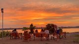 People on benches watch a sunset over water. Orange and yellow sky; a solar panel stands nearby.