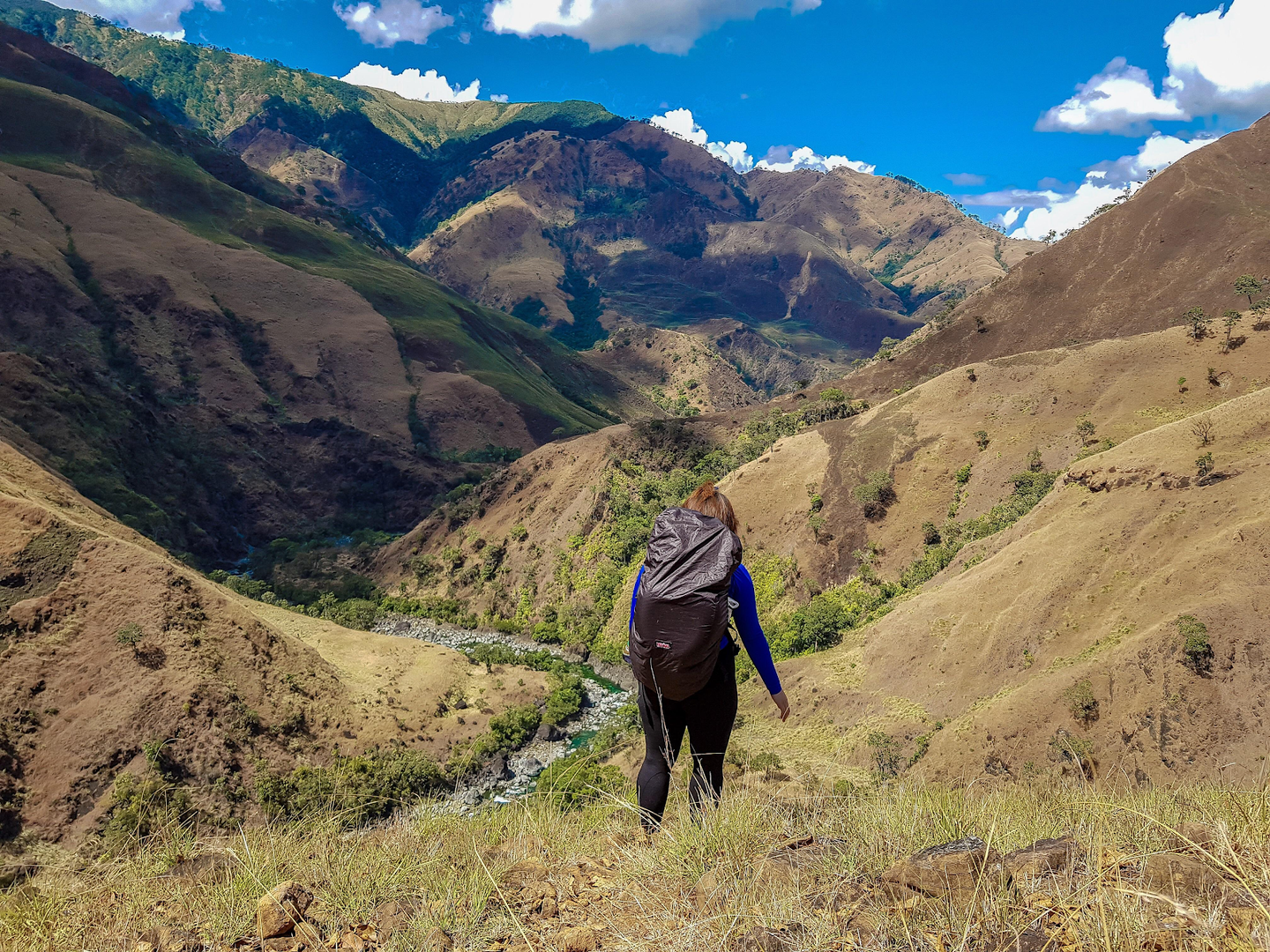 Person backpacking down a grassy mountain slope toward a valley. Mountains rise on either side under a blue sky.