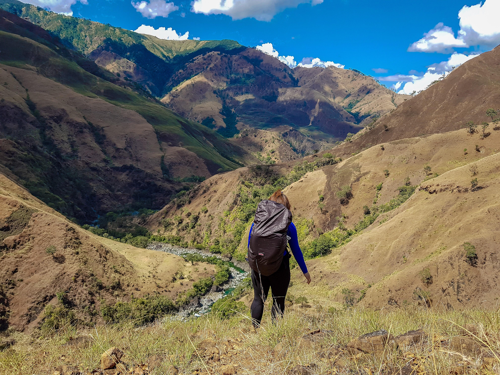 Person backpacking down a grassy mountain slope toward a valley. Mountains rise on either side under a blue sky.