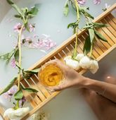 Hand holding a drink in a bathtub, with flowers and wooden tray; serene and relaxing.