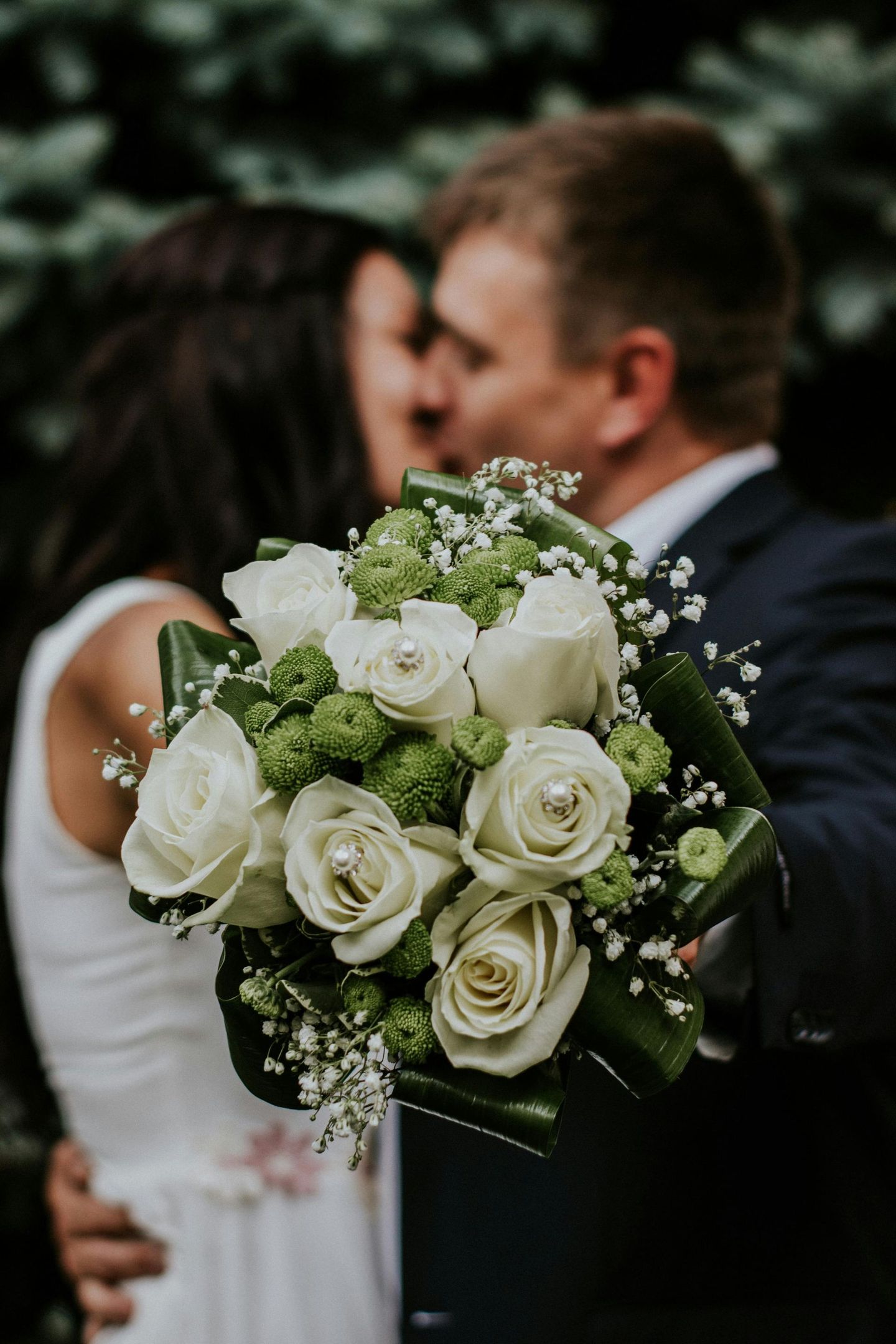 Wedding bouquet with white roses in focus, couple kissing in background.