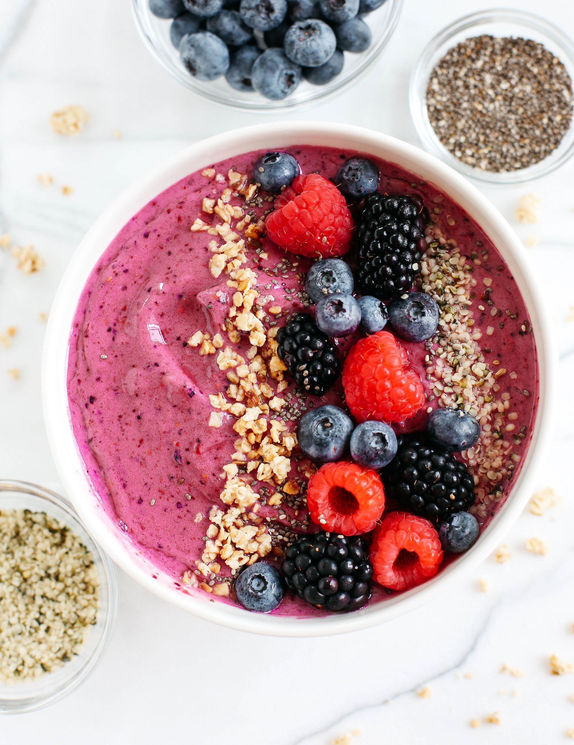 A bowl of vibrant purple berry smoothie topped with raspberries, blackberries, blueberries, chopped nuts, and hemp seeds.