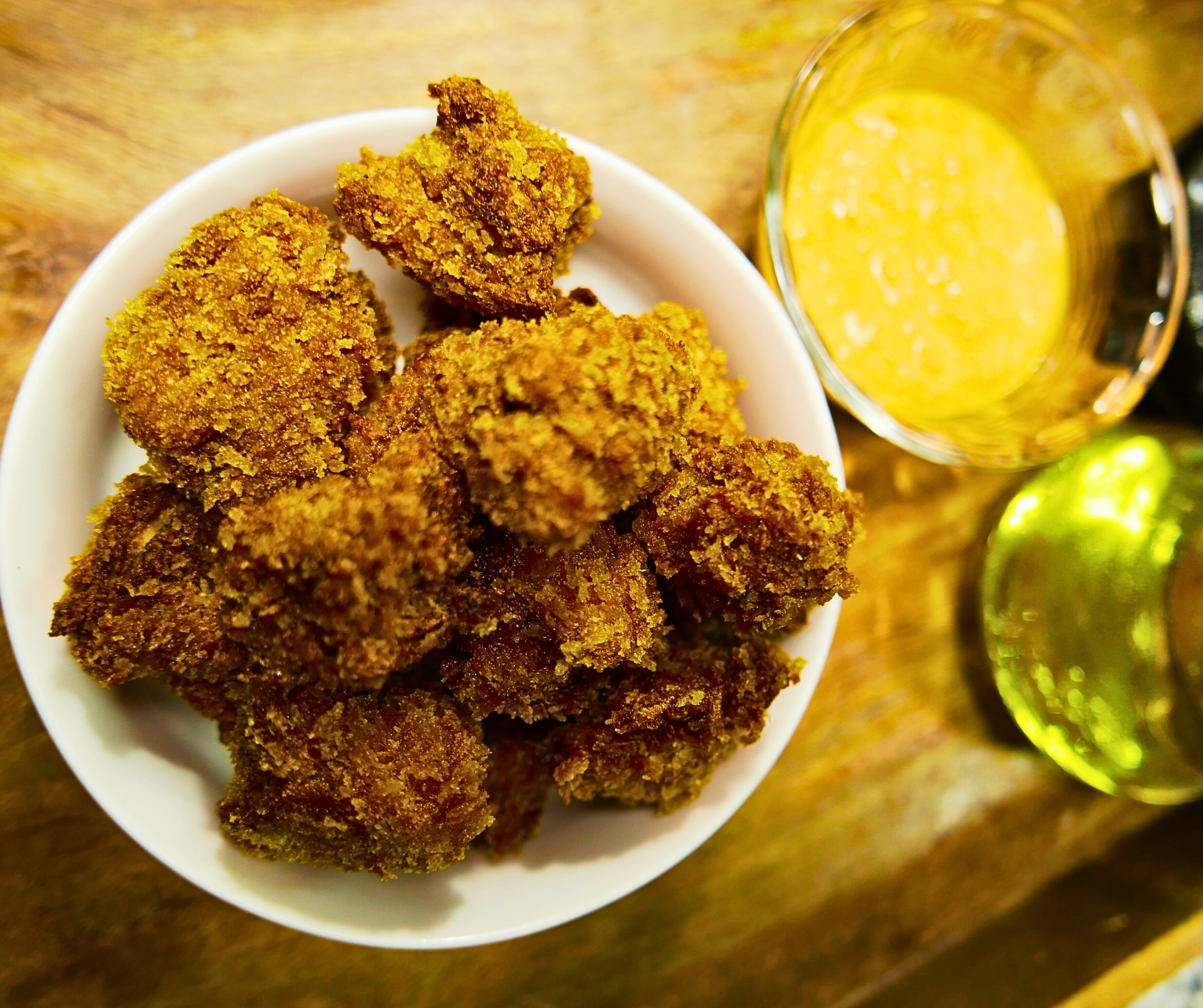Fried chicken nuggets in a white bowl with dipping sauce and olive oil on a wooden surface.