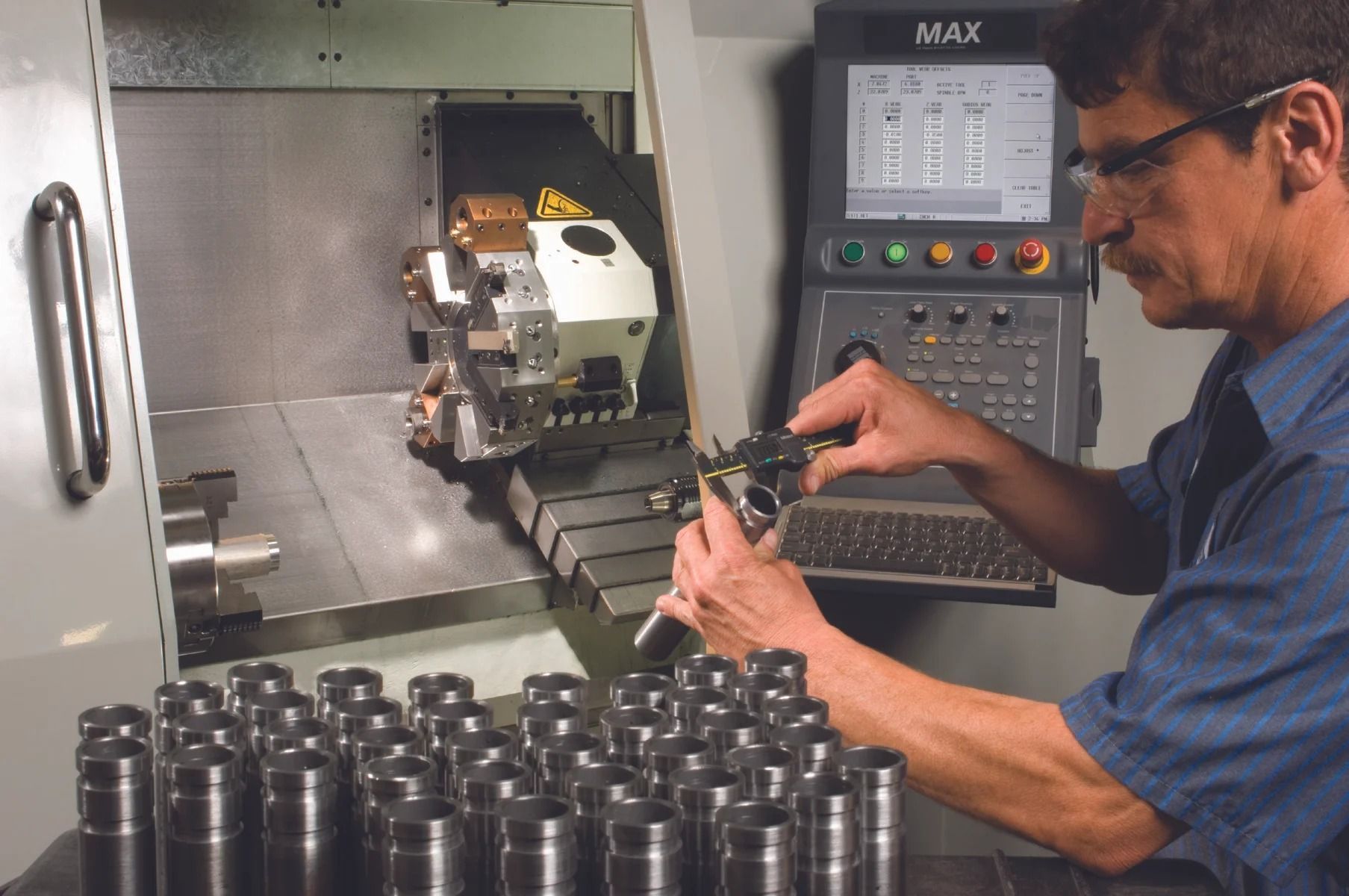 Man working at a CNC machine, inspecting a part. Machine control panel visible. Parts in foreground.