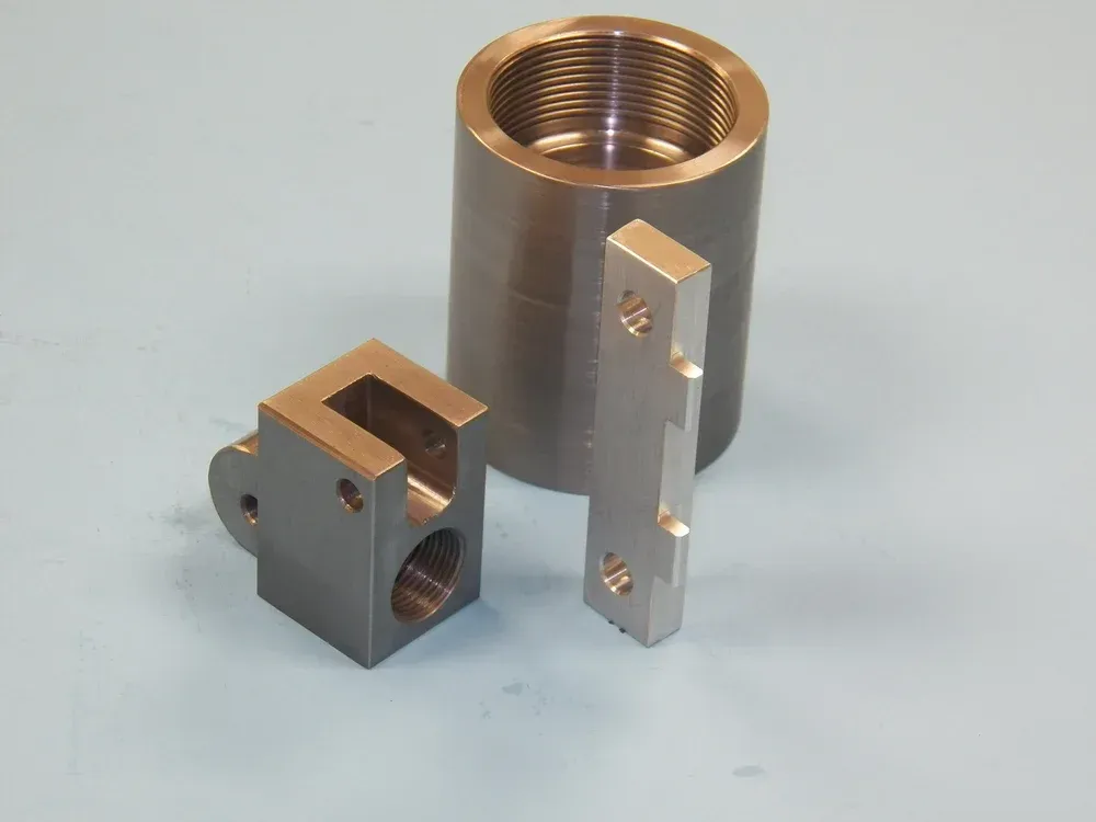 Machined metal parts: a threaded cylinder, a rectangular bar, and a block with cutouts, on a blue surface.