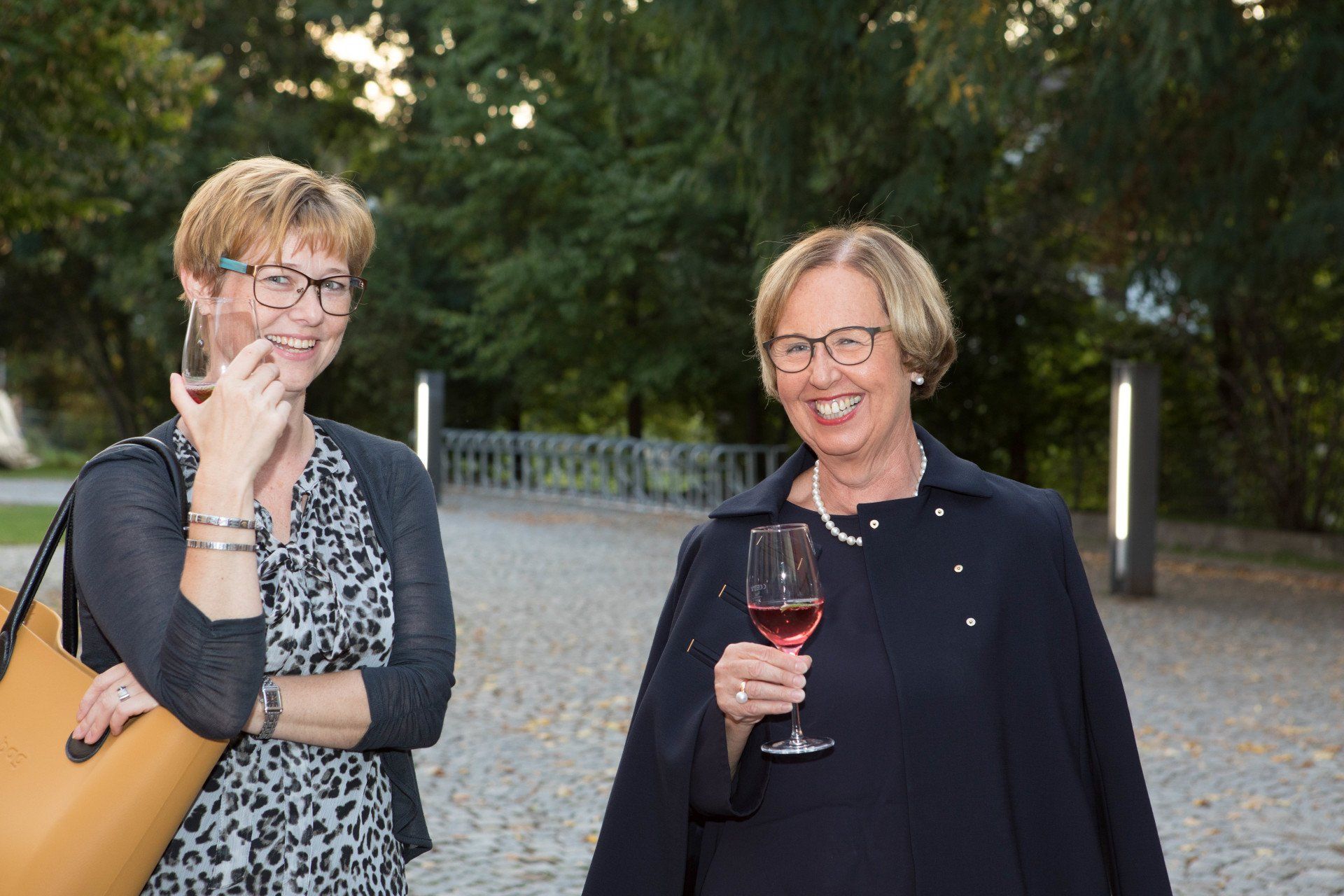 Zwei Frauen stehen nebeneinander, halten Weingläser und lächeln.