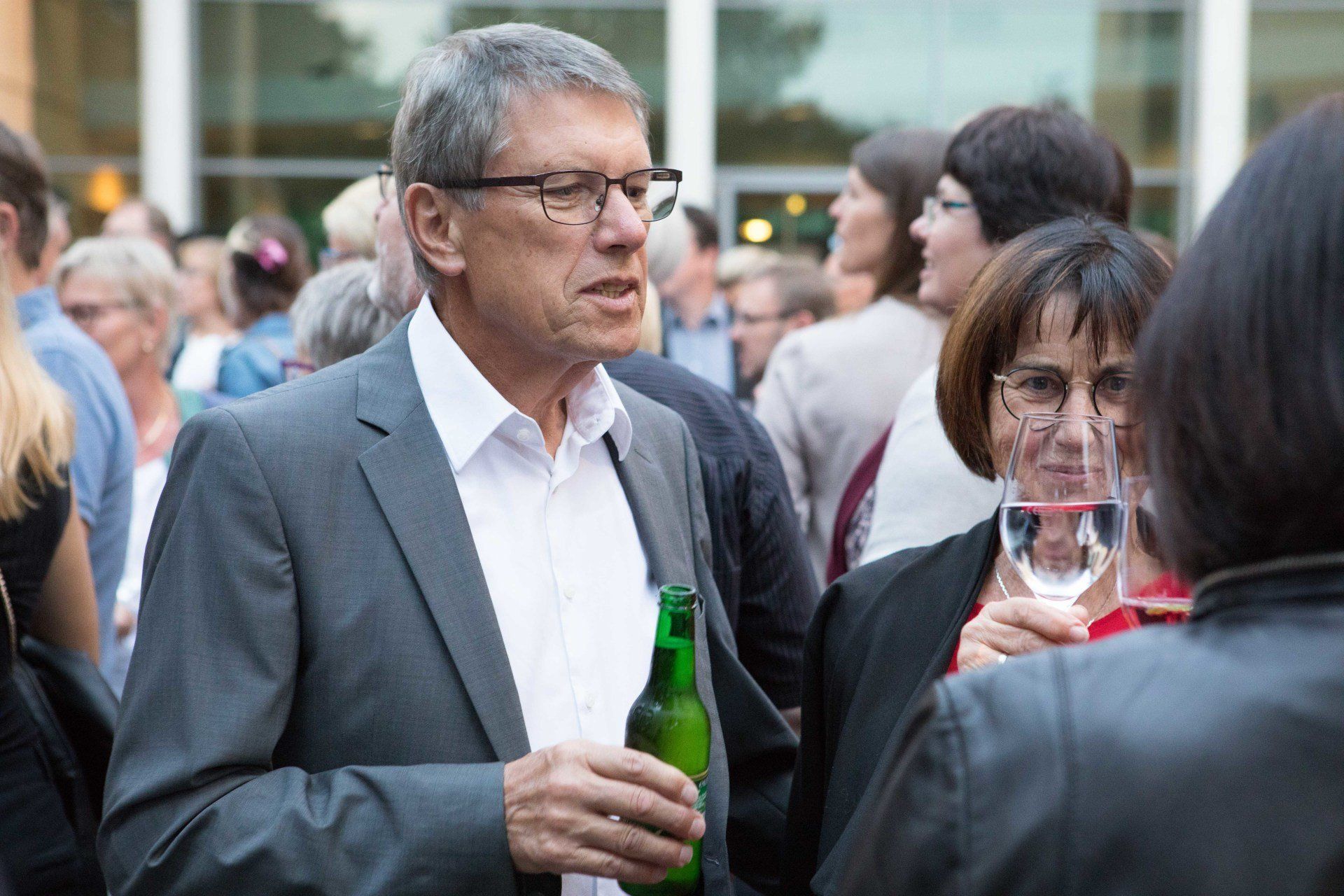 Ein Mann im Anzug hält eine Flasche Bier und ein Glas Wein in der Hand.