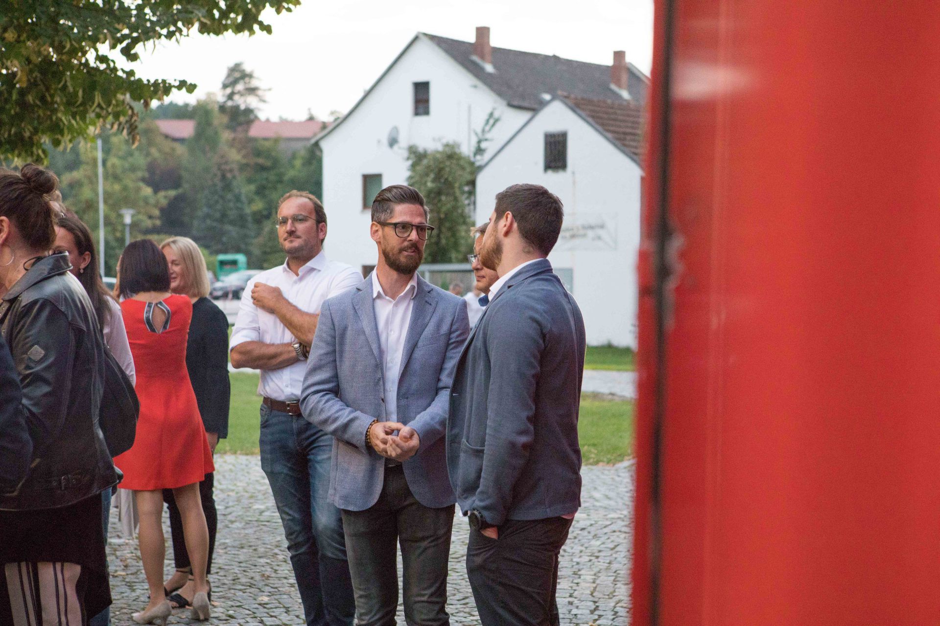 Eine Gruppe von Menschen steht vor einem roten Gebäude und unterhält sich.